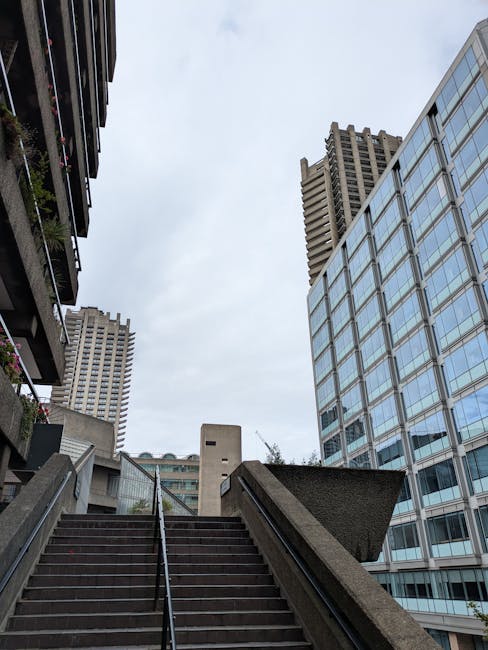 View looking up from a set of outdoor concrete stairs with metal handrails, surrounded by urban architecture. On the left side, a multi-story residential building with balconies and greenery visible o