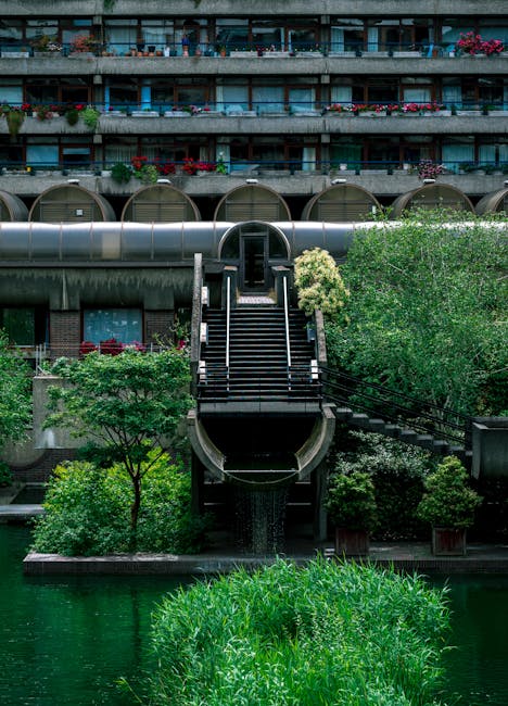 View looking up from a set of outdoor concrete stairs with metal handrails, surrounded by urban architecture. On the left side, a multi-story residential building with balconies and greenery visible o
