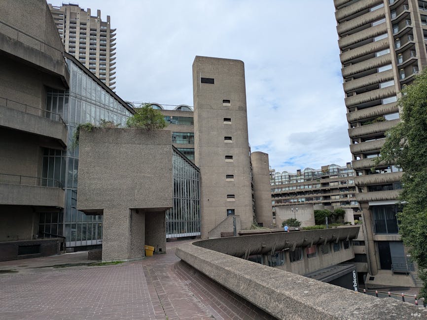 A low-angle view of a modern urban courtyard surrounded by Brutalist-style concrete buildings, with textured surfaces and geometric shapes. In the foreground, there is a curved concrete walkway with a